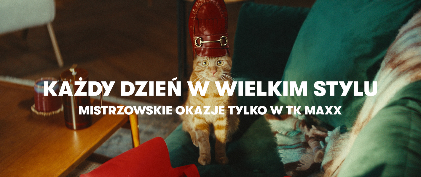 Ruda kotka z czerwonym, lśniącym mokasynem na głowie siedzi na zielonej kanapie obok czerwonej torby na zakupy z TK Maxx.